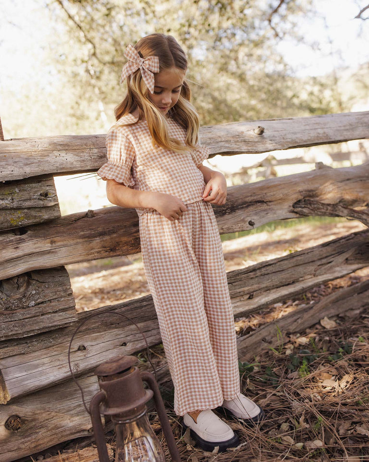 A young girl wearing the rylee + cru francie blouse in fawn gingham with a matching bow stands by a rustic wooden fence outdoors, looking down and holding her clothes, with a lantern on the ground nearby.