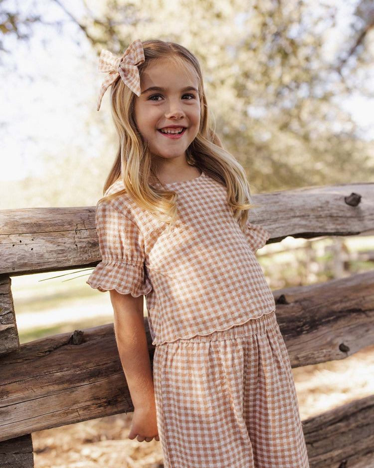 A smiling young girl in the rylee + cru francie blouse in fawn gingham, with a matching hair bow, leans against a rustic wooden fence outdoors amid sunlight and trees.