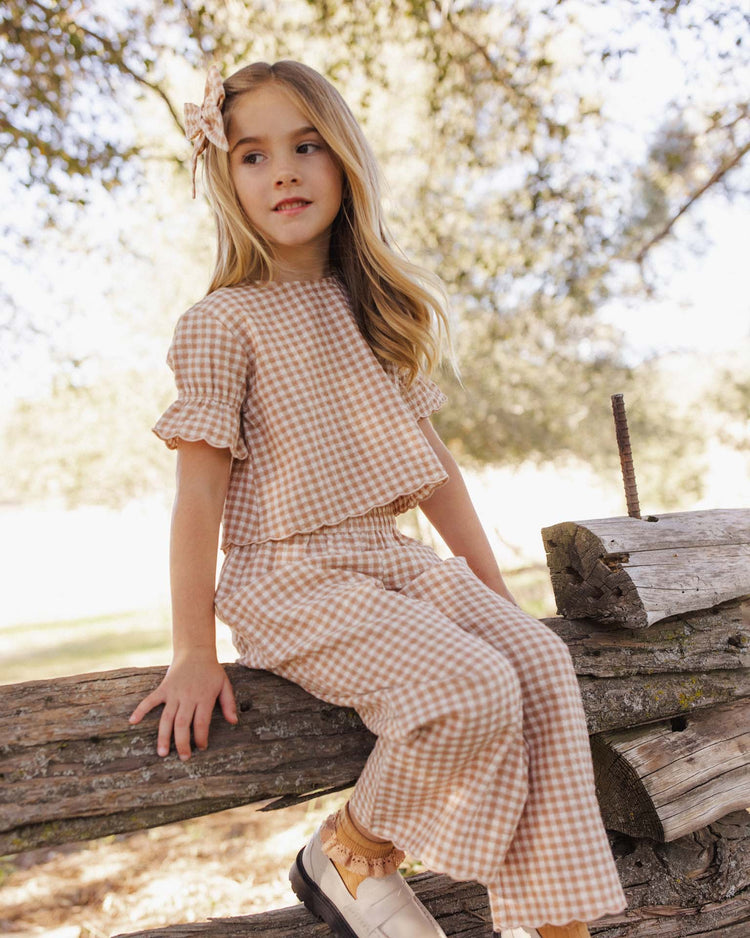 A young girl with long blonde hair sits on a log outdoors, surrounded by trees and sunlight, wearing the rylee + cru francie blouse in fawn gingham.