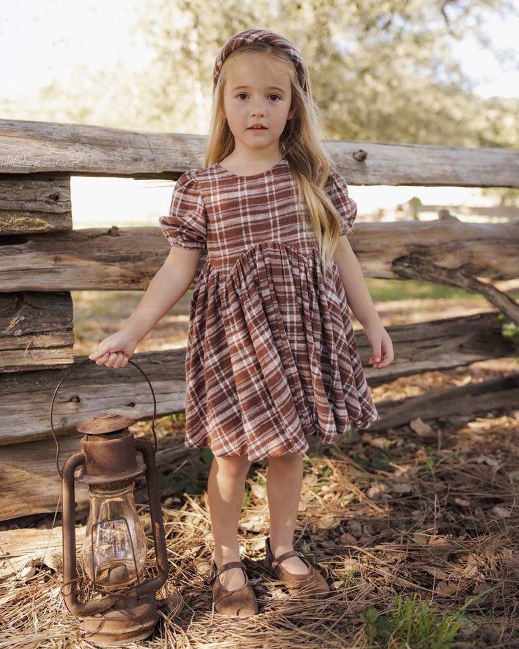 A young girl wears the rylee + cru rose dress in aspen plaid, featuring puff sleeves, as she stands outdoors by a wooden fence holding a vintage lantern. Pine needles blanket the ground and trees fill the background.
