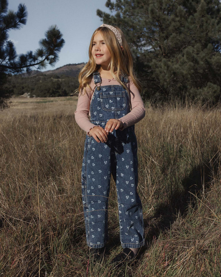 A young girl in patterned denim overalls and the rylee + cru ribbed long sleeve in rosewood, featuring a delicate lace-trimmed neckline, stands smiling in a grassy field with trees and hills in the background as she looks to the side.