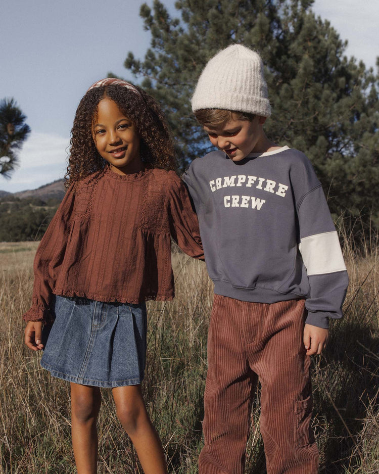 Two children stand together outdoors in a grassy field, smiling. The girl wears the cecile blouse in mulberry by rylee + cru paired with a denim skirt, while the boy wears a knit hat, blue Campfire Crew sweatshirt, and brown pants. Pine trees are in the background.