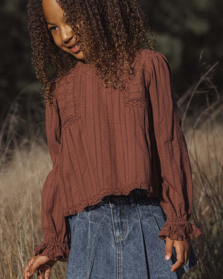 A young child with curly hair stands outdoors, smiling slightly while surrounded by tall dry grass, wearing the rylee + cru cecile blouse in mulberry paired with a denim skirt.