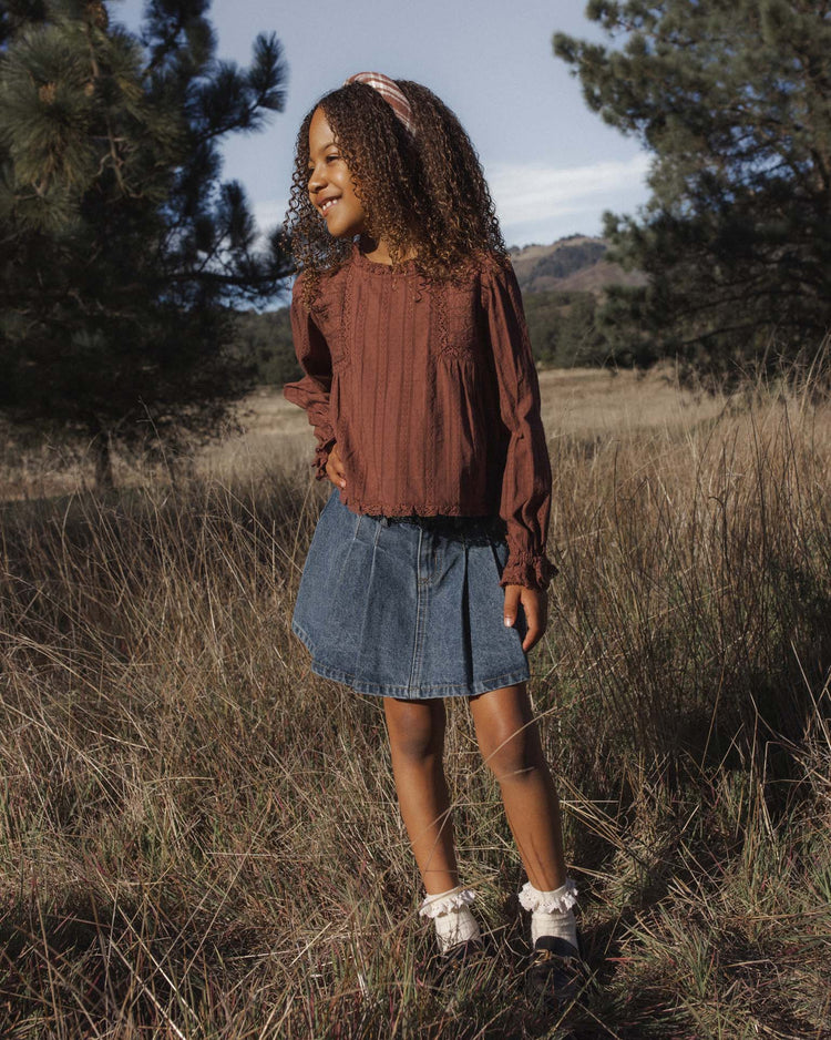 A young girl with curly hair stands smiling in a grassy field. She wears the rylee + cru cecile blouse in mulberry, paired with a denim skirt, white socks, and dark shoes. Trees and hills stretch out in the background under a clear sky.