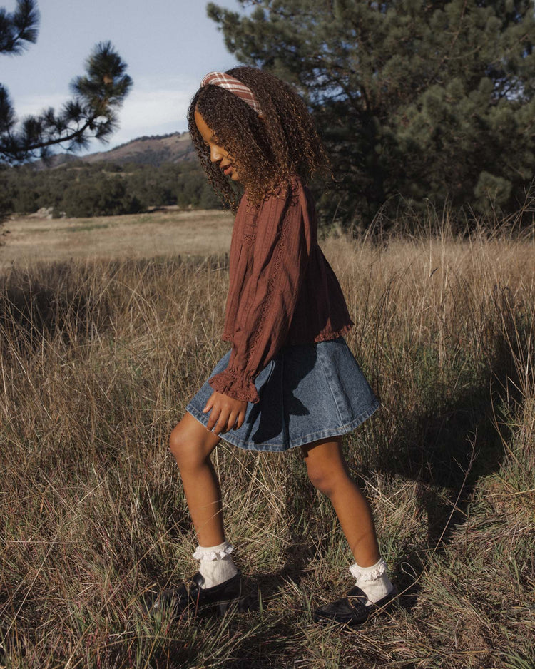 A young girl with curly hair and a headband walks through a grassy field wearing the Cecile Blouse in Mulberry by rylee + cru—a brown long-sleeve cotton prairie top with delicate lace trim—paired with a denim skirt, socks, and black shoes. Pine trees and hills are visible in the background.