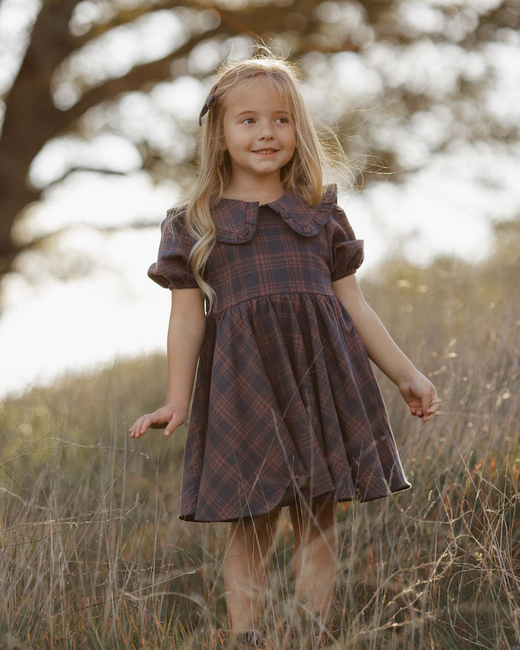 A young girl with long blonde hair stands smiling in a grassy field, wearing the rylee + cru camille dress in midnight plaid. Made from 100% cotton, the dress perfectly suits the natural setting, with trees and gentle sunlight in the background.