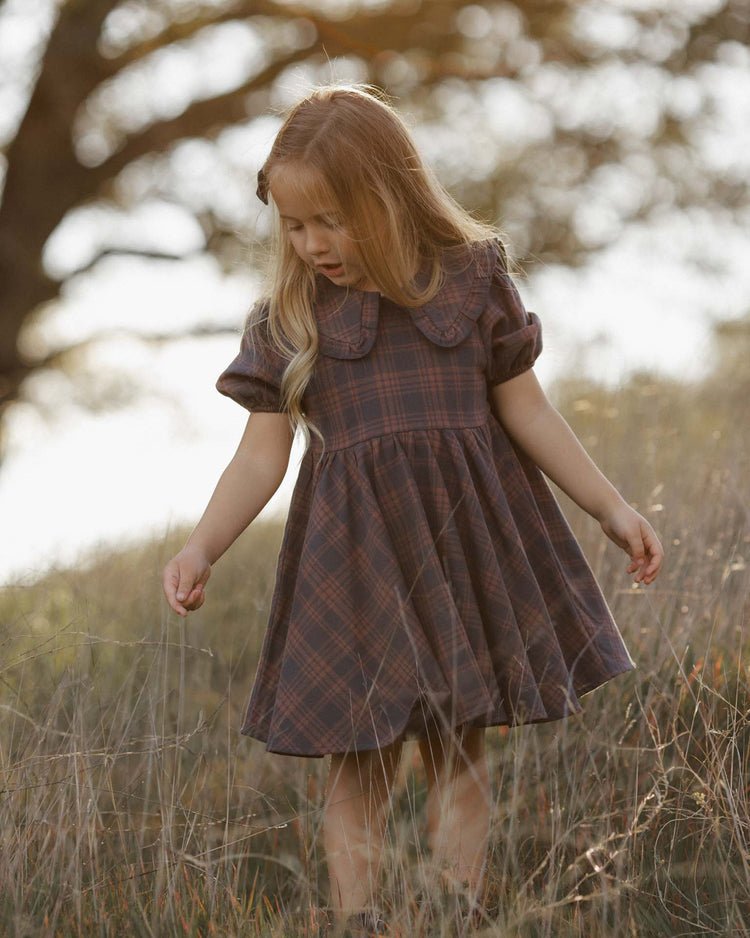 A young girl with long blond hair stands in a grassy field, looking down and gently touching the tall grass. She wears the rylee + cru camille dress in midnight plaid, crafted from 100% cotton, as sunlight filters through the trees behind her.