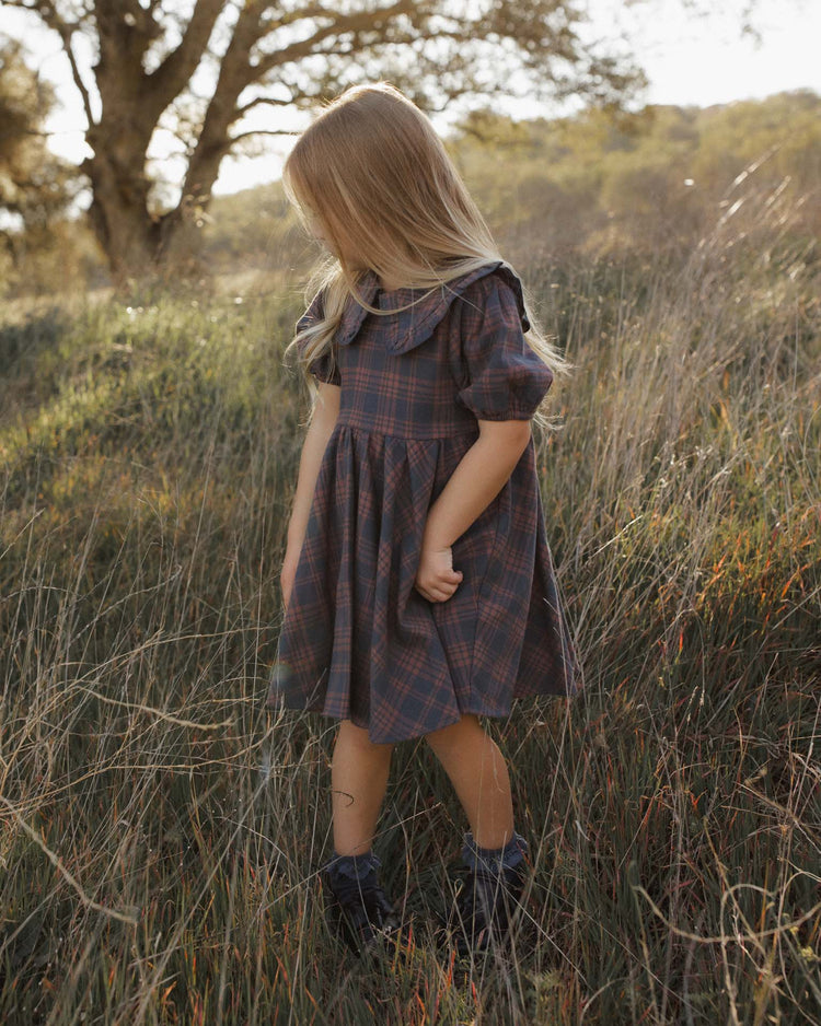 A young girl with long blonde hair stands in tall grass, looking down and to the side, wearing the rylee + cru camille dress in midnight plaid, crafted from 100% cotton, with sunlight filtering through trees in the background.