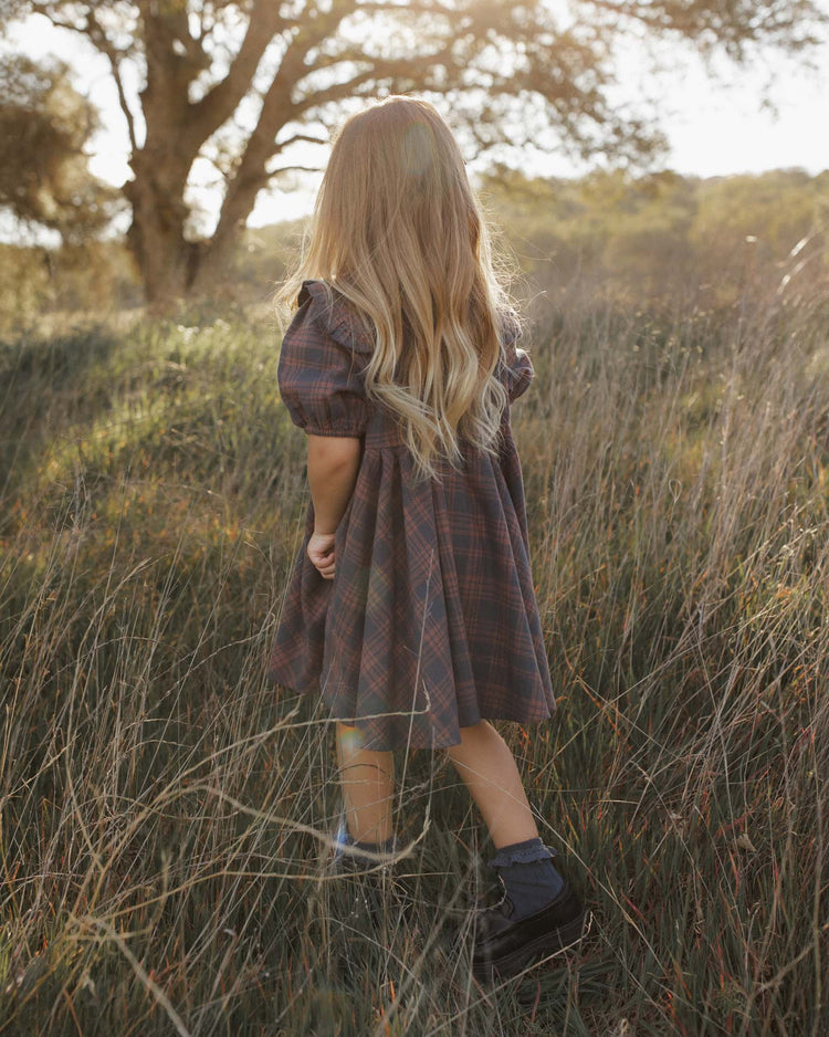 A young girl with long blond hair stands in tall grass, facing away, sunlight streaming through the trees as she wears the camille dress in midnight plaid by rylee + cru. Crafted from 100% cotton, the mini dress moves gently in the breeze while golden light filters over the peaceful scene.