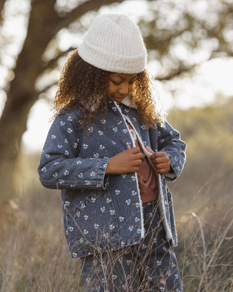A child with curly hair wears a cream knit beanie and the rylee + cru long chore coat in heart ditsy — a blue floral autumn-winter jacket featuring a cosy shearling lining — standing outdoors on grassy ground as sunlight filters through the trees.
