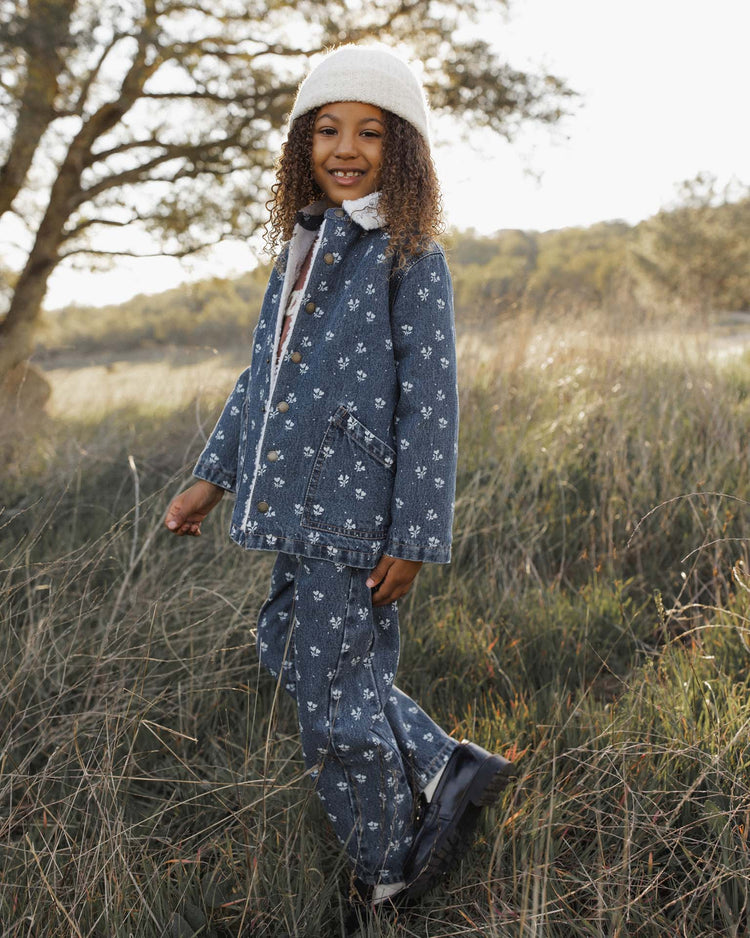 A smiling child stands in a grassy field, wearing the rylee + cru long chore coat in heart ditsy—a cozy autumn-winter piece with soft shearling lining for extra warmth—along with a white knit hat and coordinating pants, framed by trees and sunlight in the background.