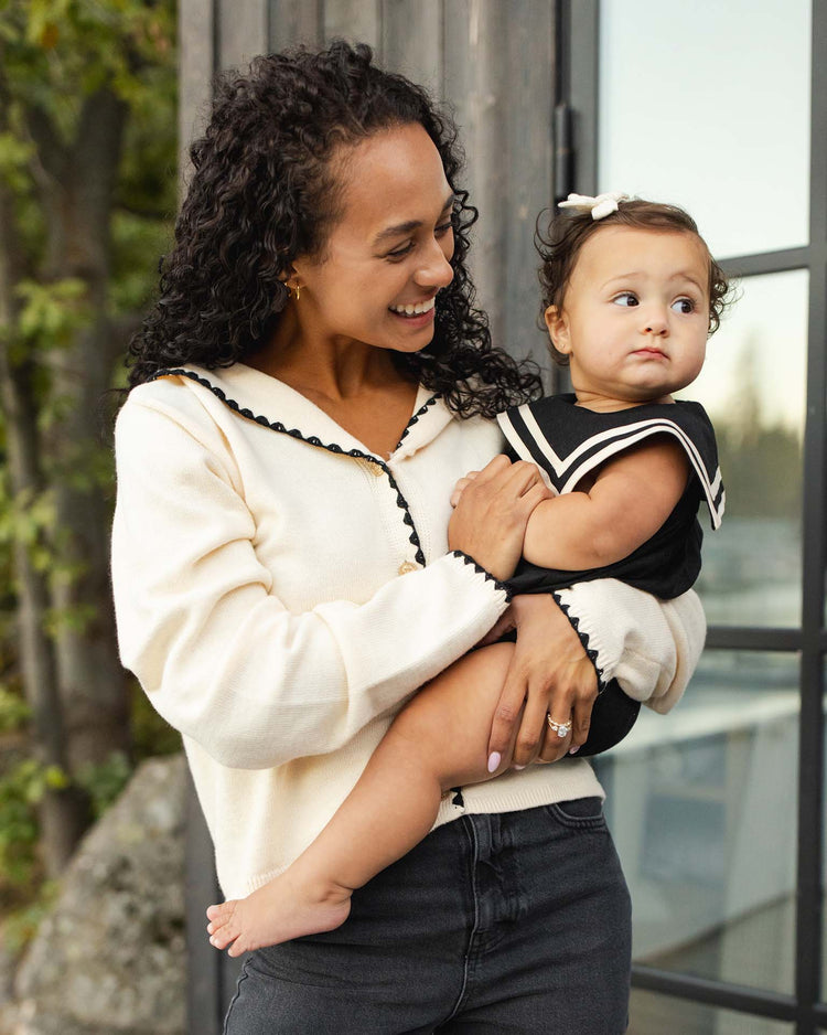 A woman with curly hair smiles while holding a baby girl dressed in the rylee + cru sailor romper in black, standing outdoors near a glass door with greenery in the background.