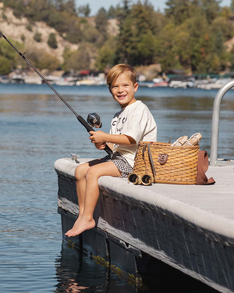 A young boy wears the rylee + cru relaxed tee in catch the wave, sitting barefoot on a lake dock with a fishing rod, picnic basket, and towels beside him. Trees and boats can be seen in the background as he smiles.