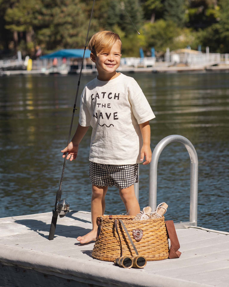 A young boy stands barefoot on a dock, smiling in the rylee + cru relaxed tee in catch the wave, paired with checkered shorts. A wicker basket and toy binoculars sit nearby, with a lake and trees behind him.
