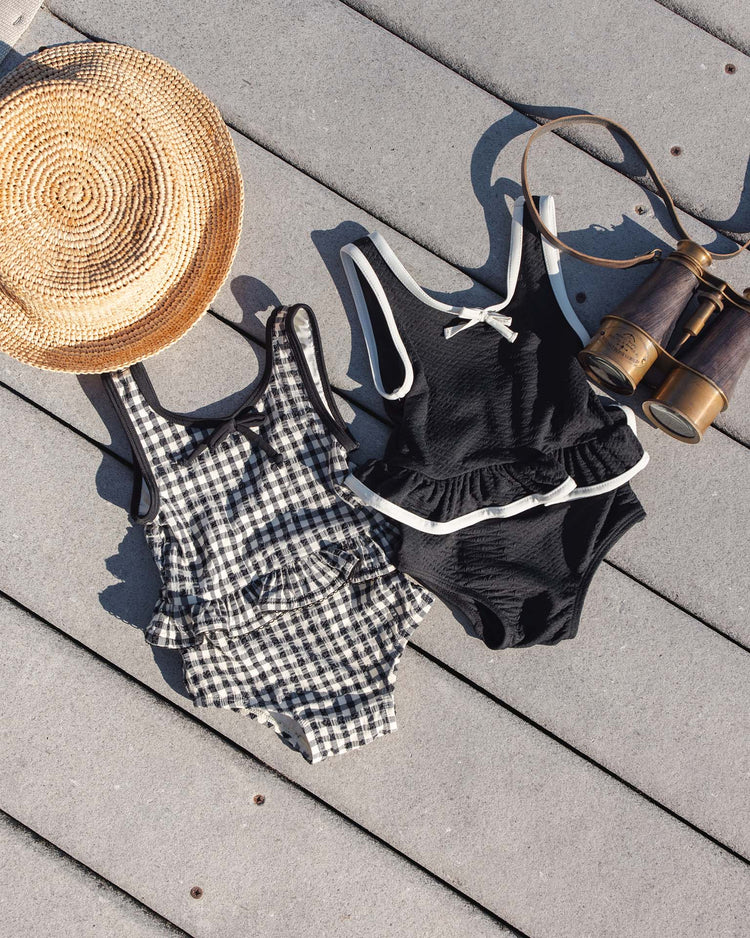 A rylee + cru skirted one-piece in black gingham lies on a wooden deck beside a straw sunhat and vintage binoculars as sunlight casts soft shadows across this charming UPF 50+ scene.