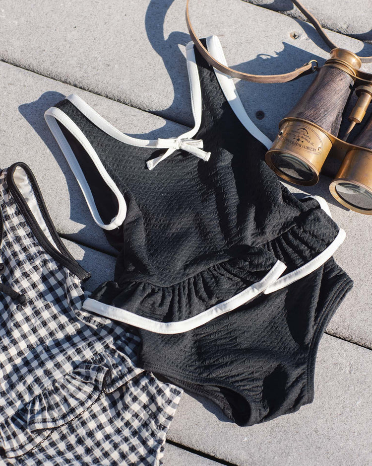 The rylee + cru skirted one-piece in black, featuring white trim and UPF 50+ protection, is displayed on a light stone surface beside black-and-white gingham swim bottoms and vintage binoculars with a leather strap.