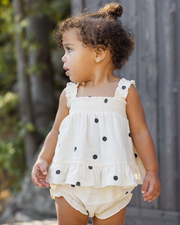 A young child wearing the rylee + cru lala set in polka dot stands outdoors, looking to the side, with trees and a wooden fence in the background.