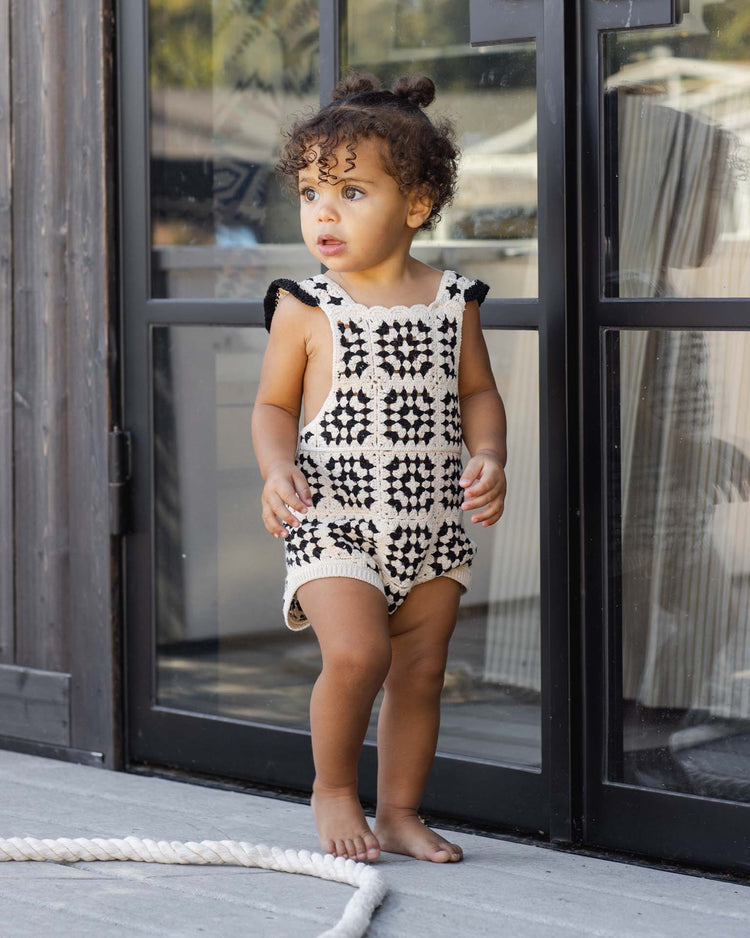 A toddler with curly hair stands barefoot outside a glass door, wearing the rylee + cru crochet romper in abstract, adding vintage charm. The child looks to the side with curiosity as a thick white rope rests nearby.