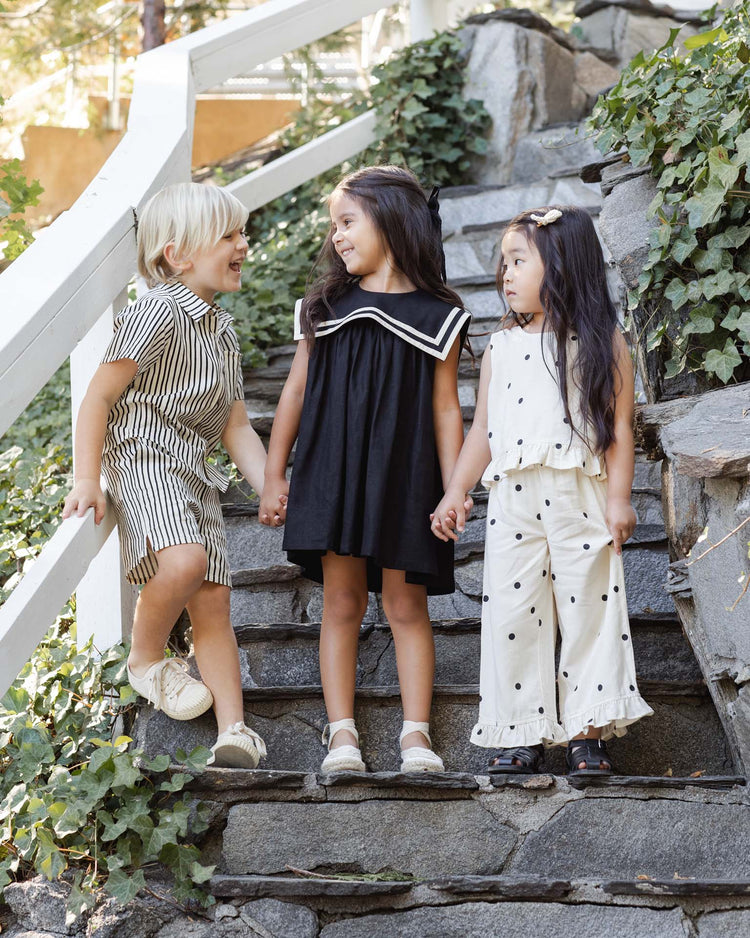 Three kids stand on stone steps, smiling amid greenery. The boy wears the rylee + cru collared short sleeve shirt in wavy stripe, the center girl wears a black dress, and another girl is in a cream outfit with black polka dots.