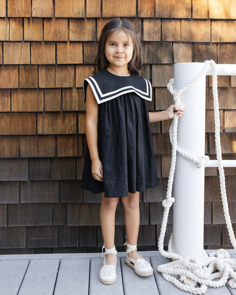 A young girl wears the rylee + cru sailor dress in black, featuring a white-trimmed collar, as she stands on a wooden deck holding a rope-wrapped post with brown shingle wall in the background.