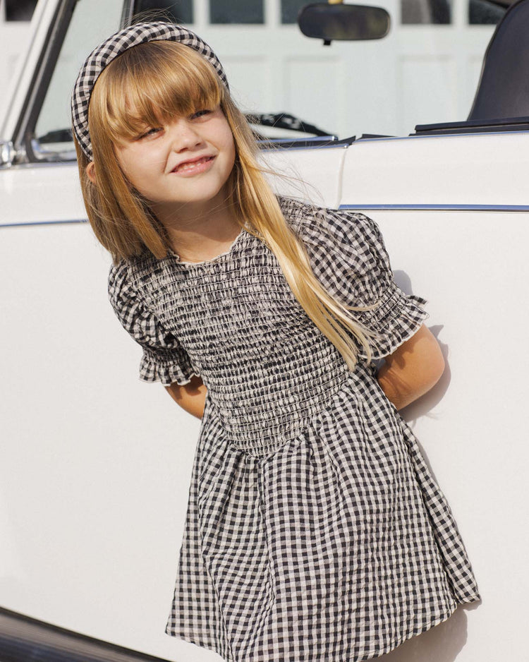 A young girl with long blonde hair smiles outdoors beside a white car, wearing a black-and-white checkered dress and the rylee + cru padded headband in black check, with her hands behind her back.
