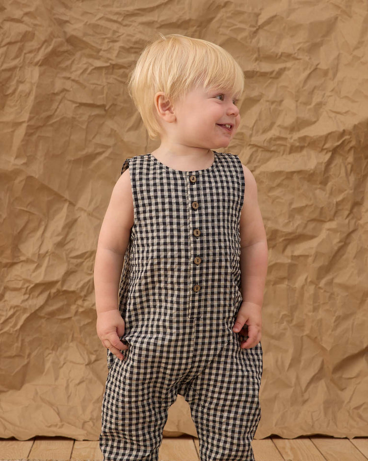 A smiling blonde toddler stands on a wooden floor, wearing the rylee + cru button jumpsuit in black check, with a crumpled brown paper backdrop behind.