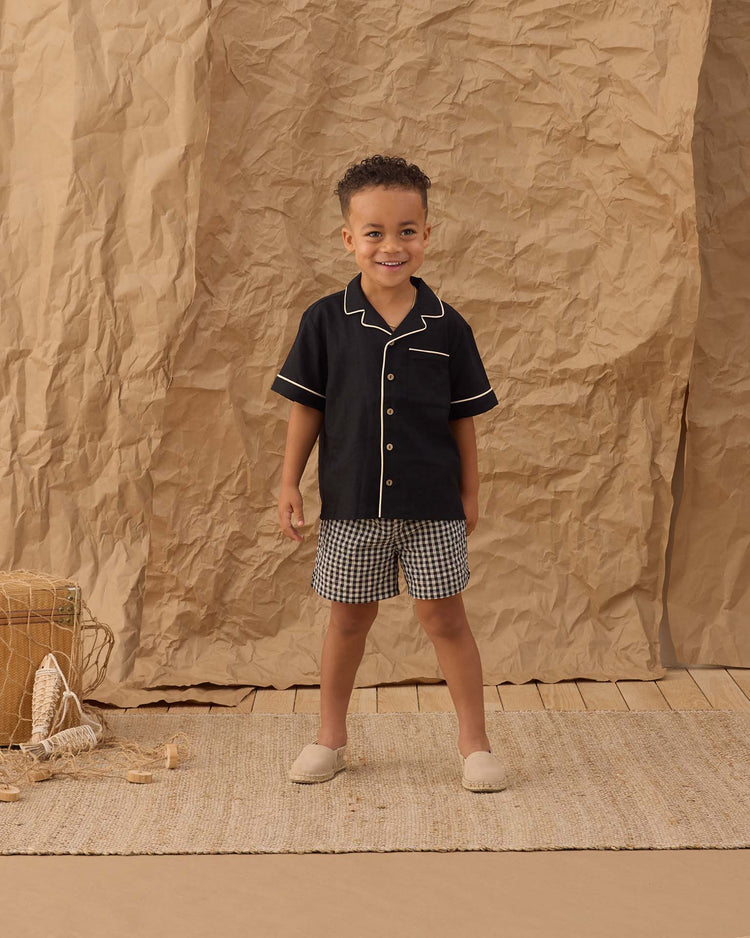 A young child smiles on a woven rug, wearing rylee + cru's perry short in black check with a black piped shirt, beige slip-on shoes, and a crumpled brown paper backdrop for a vintage-inspired look.