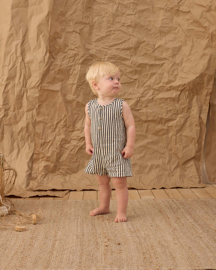 A young child with blond hair stands barefoot on a woven rug, wearing the rylee + cru mavrick romper in wavy stripe. Against a backdrop of crumpled brown paper and wood flooring, the child looks to the side.