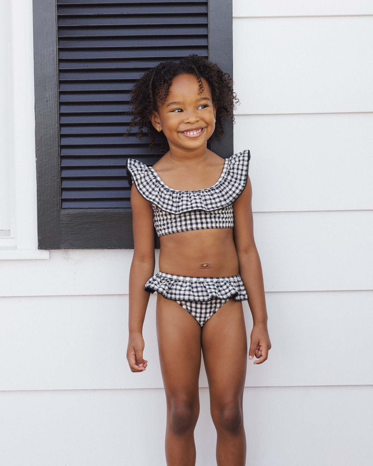 A young girl stands smiling before a white wall with black shutters, wearing the rylee + cru margot bikini in black gingham.