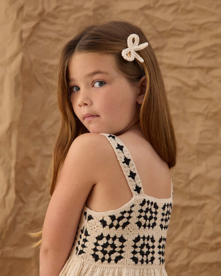 A young girl with long brown hair wears a rylee + cru crochet bow clip in ivory, looking over her shoulder against a crumpled brown paper background.