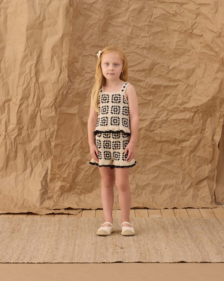 A young girl with long blonde hair stands on a woven rug, wearing the kira set in abstract by rylee + cru. The background is crumpled brown paper, and she gazes at the camera with a neutral expression.
