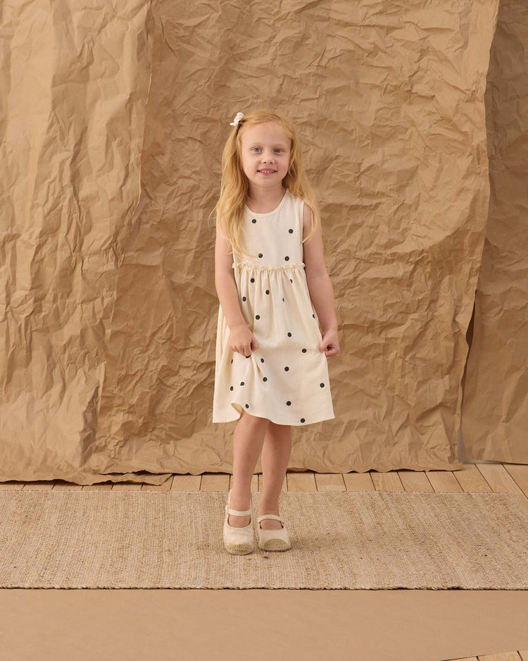 A young girl with long blonde hair smiles in front of a textured brown backdrop, wearing the rylee + cru harper dress in polka dot with coordinating bloomers.