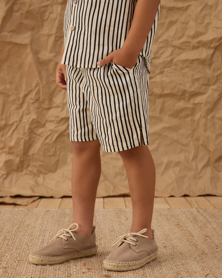 A child stands with one hand in their pocket, wearing rylee + cru’s perry short in wavy stripe and a matching shirt for a vintage look. They wear beige espadrilles, set on textured brown paper and a woven natural fiber floor.