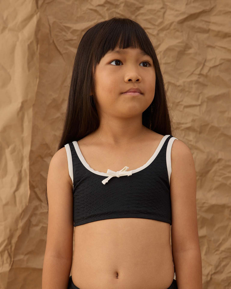 A young girl with long, straight hair wears the rylee + cru daisy bikini in black, featuring white trim and a small white bow made from recycled polyamide, standing before a crumpled brown paper background.