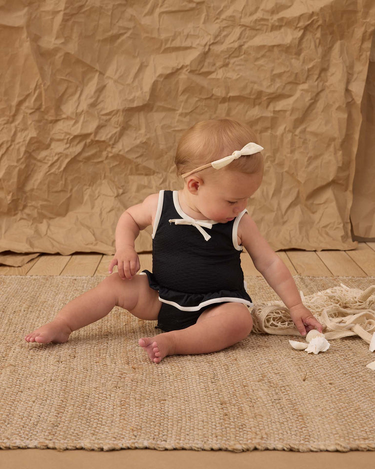 A baby in a rylee + cru skirted one-piece in black with white trim and a white headband sits on a woven rug, reaching for a white object against a textured brown backdrop.