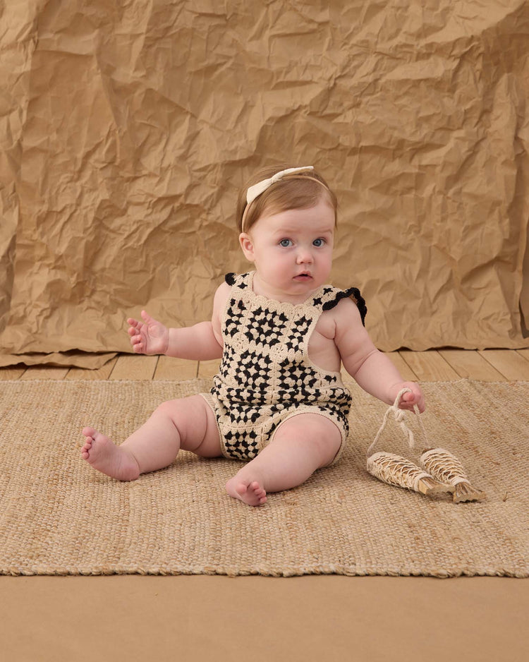 A light-skinned, blue-eyed baby sits on a woven rug, wearing the rylee + cru crochet romper in abstract (100% cotton) and a cream headband, holding a small woven toy. Crumpled brown paper adds vintage charm to the background.