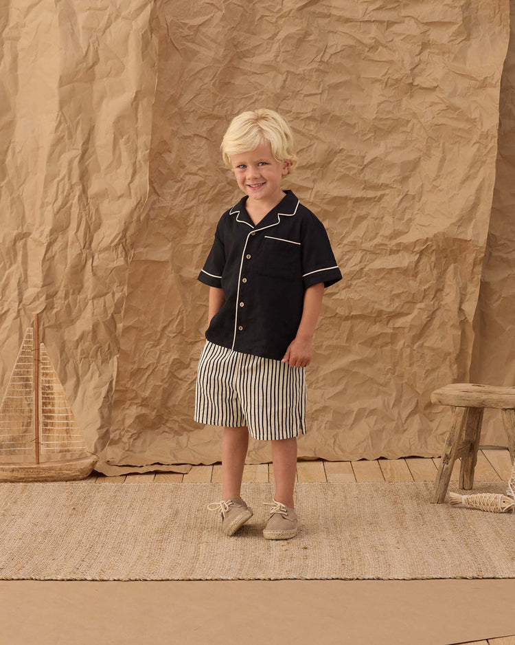 A young boy with blond hair stands on a woven rug, smiling in front of a crumpled brown paper backdrop. He wears the rylee + cru lapel collared shirt in black, paired with striped shorts, beside a wooden stool and model sailboat.