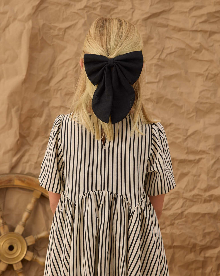 A young girl with blonde hair wears the rylee + cru oversized bow in black and a cream dress with black vertical stripes, standing before a crumpled brown paper backdrop with part of a wooden ship wheel visible on the left.