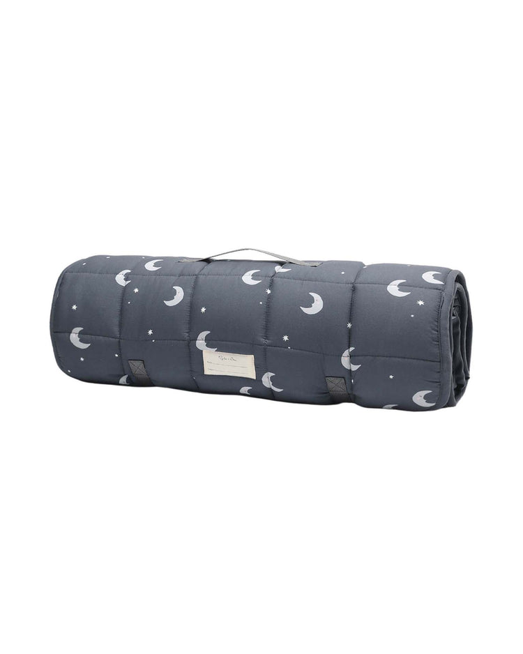 A rolled-up rylee + cru nap mat in moons, in dark gray with a moon and star pattern, featuring a carrying handle, removable pillow, micro-fleece blanket, and a front label.