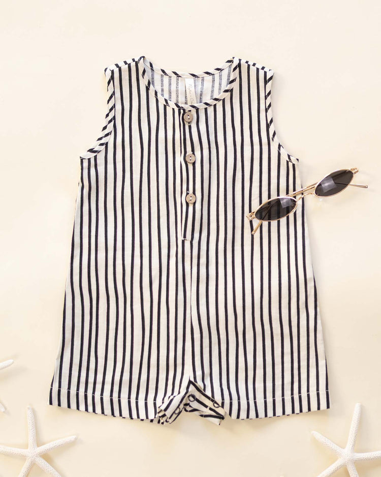 The mavrick romper in wavy stripe by rylee + cru, a sleeveless linen-viscose piece with black-and-white vertical stripes and front buttons, is styled flat beside black sunglasses and white starfish on a beige background.