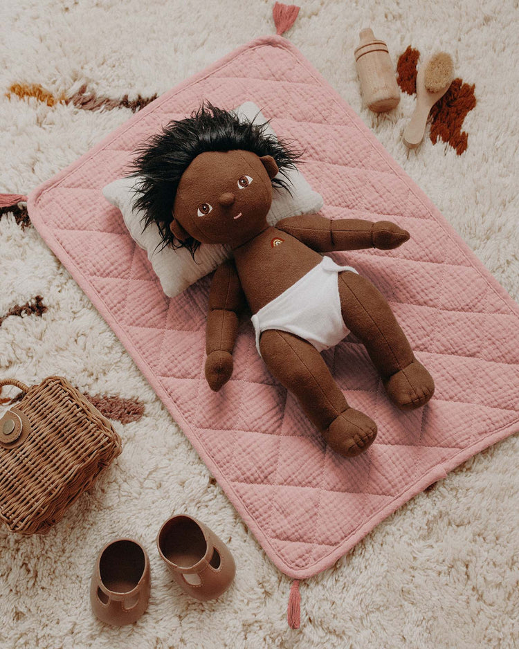 A brown plush doll with black hair, in a white diaper, lies on the Dinkum Dolls Strolley Bedding Set in Rose with a pillow, surrounded by toy accessories, baby shoes, a basket, and brushes on a patterned carpet.