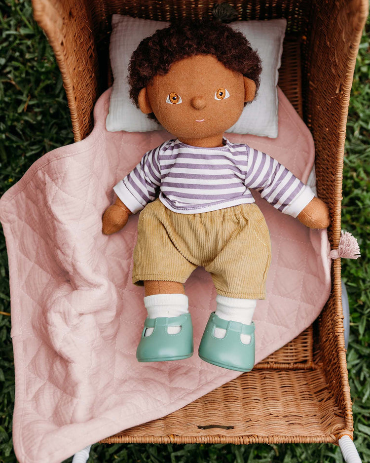 A brown-skinned cloth doll with curly hair, in a striped shirt and yellow shorts, rests on the Dinkum Dolls strolley bedding set in rose inside a wicker basket on grass.