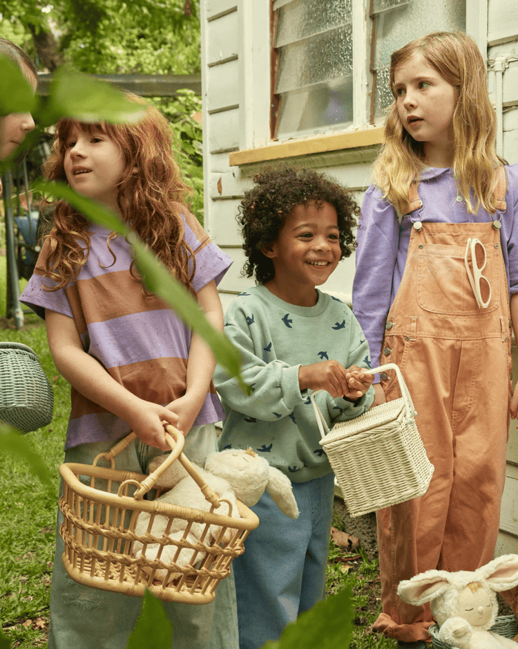 Three children stand outside a house, smiling and holding dinkum dolls tarry baskets in wheat. One has red hair and a striped shirt, another wears a bird-print sweater, and the third is in overalls. Stuffed bunnies rest on the grass.