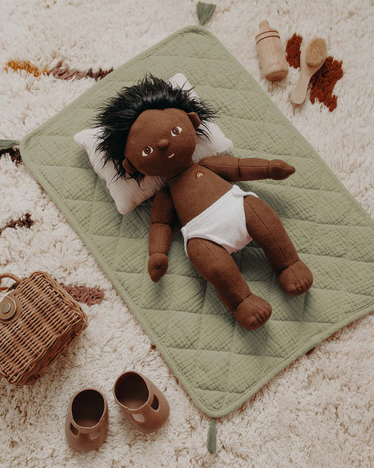 A dark-skinned Dinkum Doll with black hair, brown eyes, and a white diaper lies on a sage Strolley Bedding Set by Dinkum Dolls. Nearby are a wicker basket, brown shoes, wooden bottle, and brush on a textured rug.