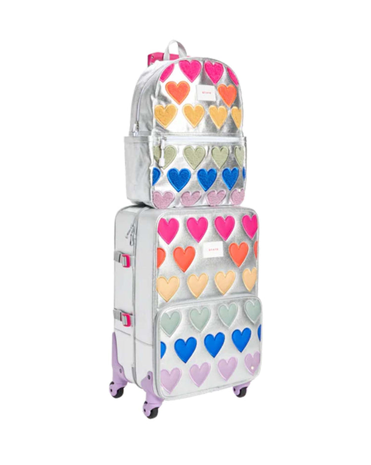 A silver "Kane Backpack in Happy Fuzzy Hearts" by State Bags is paired with a matching suitcase, both featuring vibrant heart patterns. The suitcase is accented with purple wheels and coordinating hardware. Inspired by the playful aesthetic of State Bags, this shiny, metallic-finished set is neatly stacked together.