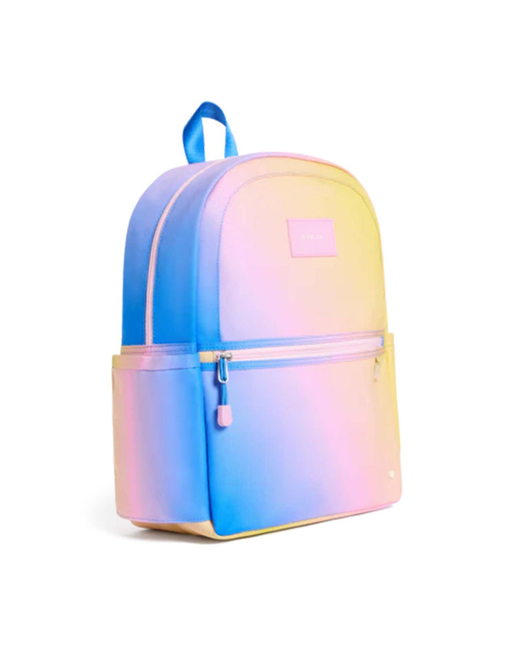 The kane backpack in blue + pink + yellow gradient by state bags is shown against a white background, featuring a pastel gradient design, a front zipper pocket, side pockets, and a blue top handle.