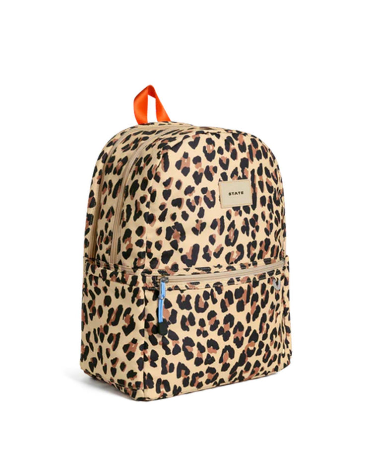 A stylish and practical kids school backpack from STATE Bags, the Kane Double Pocket Backpack in Leopard features a beige background with a bold black and brown leopard print, dual front zipper pockets, an orange top handle, and the signature STATE brand label displayed on the front.