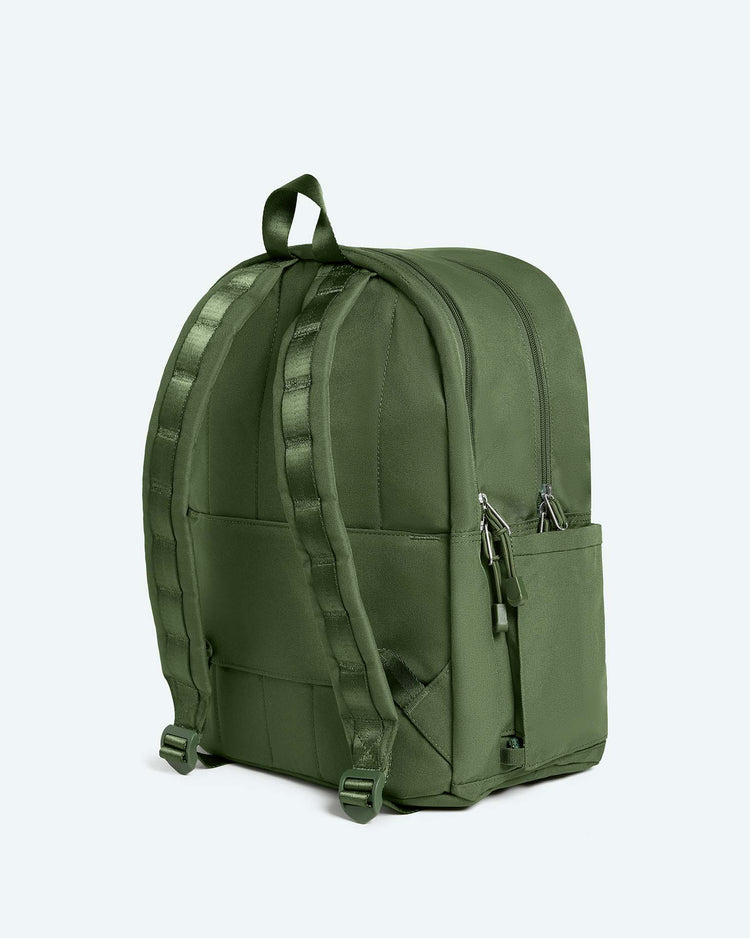 A Kane Large Double Pocket Backpack in Olive from State Bags, featuring two zippered compartments, a side pocket, and padded shoulder straps, is displayed against a light background. Some text appears partially visible but is blurred and unreadable.