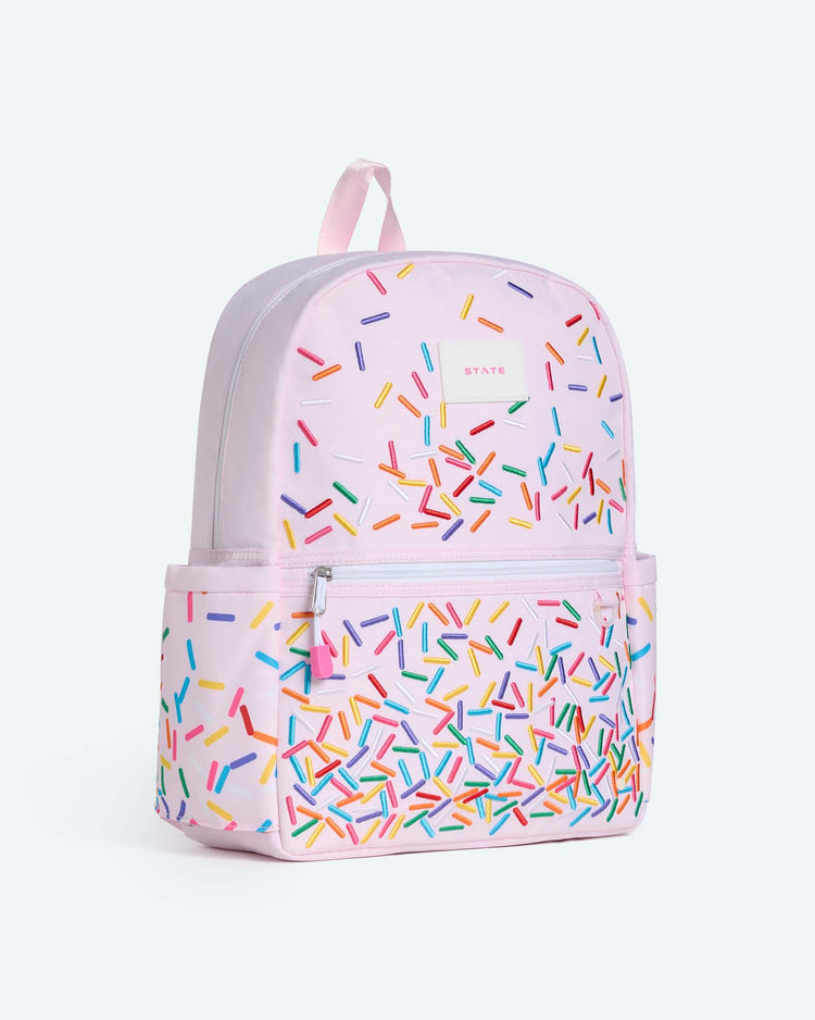 The Kane Backpack in Sprinkles by State Bags is a light pink travel backpack adorned with colorful sprinkle-like patterns. It features a front zipper pocket, side pockets, and a top handle, making it a stylish choice for school. Displayed against a plain light background.