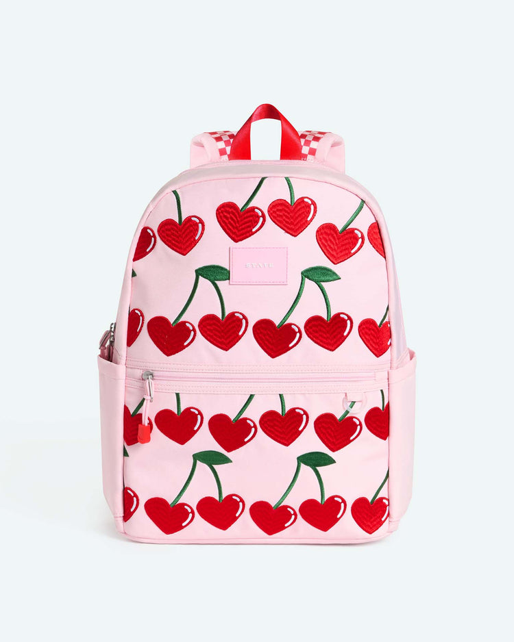 Introducing the Kane Double Pocket Backpack in Cherry by STATE Bags—a stylish pink backpack adorned with a delightful pattern of heart-shaped red cherries, green stems, and playful pink side pockets. The school backpack is finished with eye-catching red and white checkered straps for an extra dose of fun.